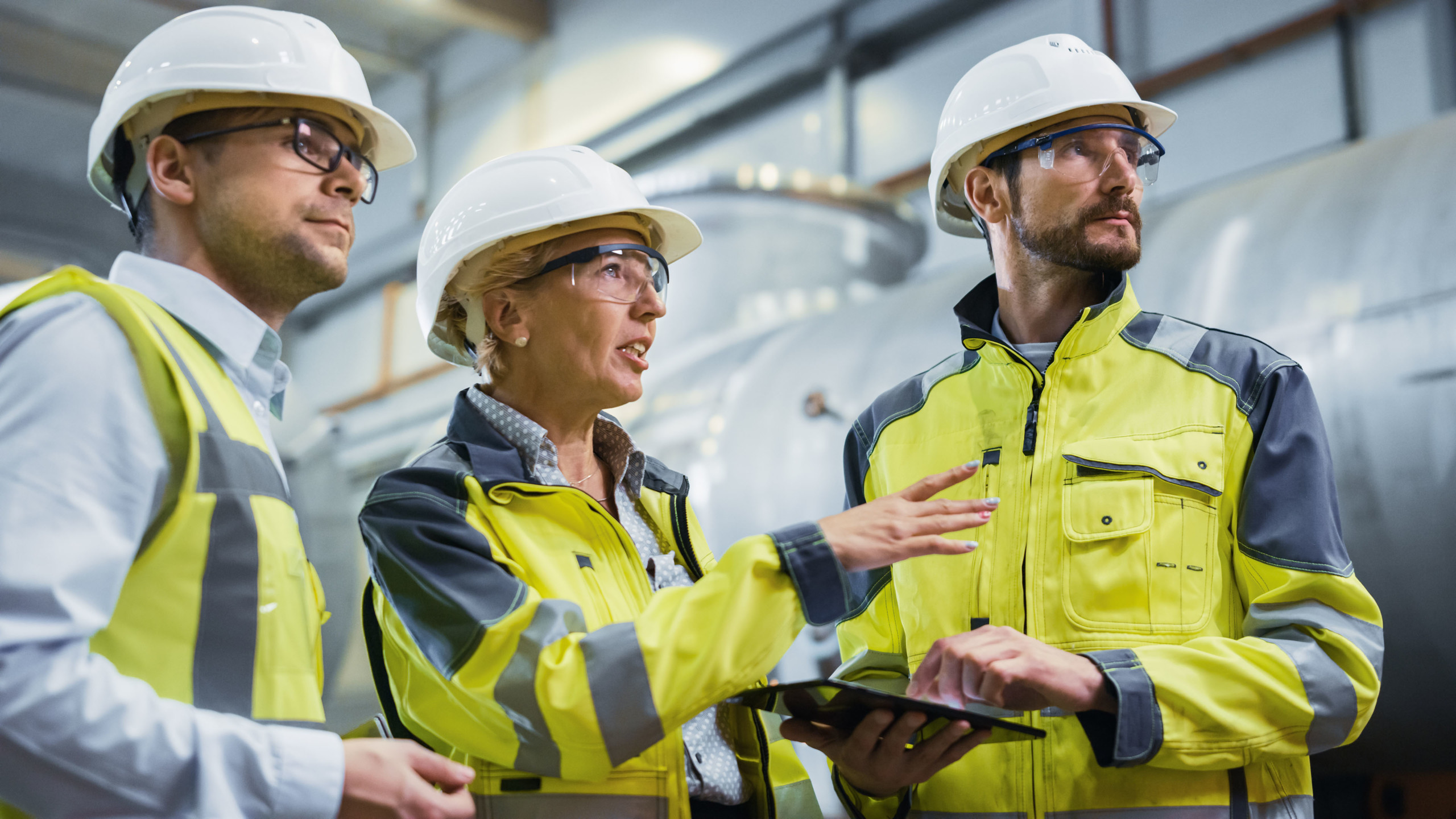 Three Heavy Industry Engineers Stand in Pipe Manufacturing Factory, Use Digital Tablet Computer, Have Discussion. Large Pipe Assembled. Design and Construction of Oil, Gas and Fuels Transport Pipeline